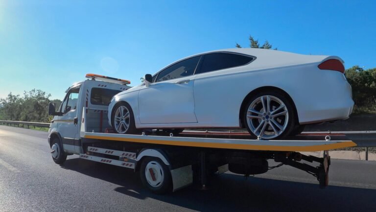 A white car being transported on the back of a flatbed tow truck on a sunny highway.