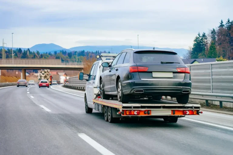 A flatbed tow truck transporting a dark gray station wagon on a multilane highway.